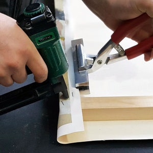 May include: A person uses a green staple gun and a red clamp to secure a canvas to a wooden frame.
