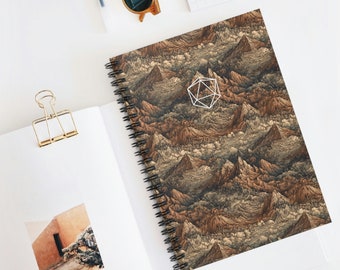 May include: A spiral-bound notebook with a detailed mountain range design in shades of brown, tan, and gray. A white geometric shape rests on the cover. The notebook is open to a blank page, with a gold paper clip and sunglasses nearby.