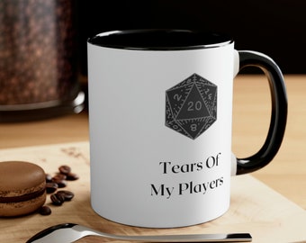 Op de afbeelding: Een witte keramische koffiemok met een zwarte rand en handvat. De mok heeft een zwart-witte illustratie van een twintigzijdige dobbelsteen en de tekst "Tears Of My Players".