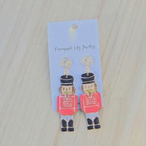Puede incluir: Un par de pendientes festivos con un diseño de soldado cascanueces. Los pendientes tienen una chaqueta roja, sombrero negro y botas negras. La tarjeta tiene el texto "Pineapple Lily Jewelry".