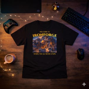 Puede incluir: Camiseta negra con el texto dorado "WELCOME TO IRONFORGE WHERE THE ALE NEVER STOPS". La camiseta presenta un gráfico detallado de una ciudad de montaña iluminada por luces cálidas. También se ven un mando de juegos, un teclado y un ratón.