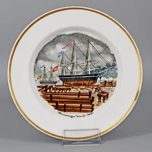 May include: White decorative plate with a gold rim, featuring a nautical scene. The image depicts a tall ship docked at a pier, with a cloudy sky backdrop. The plate has the text "Hamburger Werft 1840".