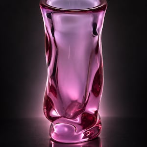 May include: A tall, cylindrical glass vase featuring a vibrant magenta and pink hue. The surface of the glass is characterized by irregular, organic indentations and a smooth, reflective finish, creating a fluid, rippled appearance throughout the body.