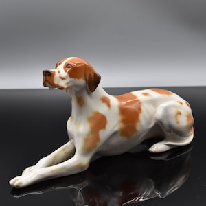 Op de afbeelding: Een keramieken hondenbeeldje in een liggende houding. De hond heeft een witte vacht met bruine vlekken en een bruine kop en oren. Het beeldje is gedetailleerd en lijkt een pointerras te zijn.