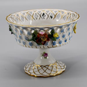 Op de afbeelding: Een witte porseleinen voetkom met een traliemotief en bloemmotieven. De kom is versierd met handgeschilderde bloemen in tinten rood, geel, blauw en groen. De kom is afgewerkt met gouden accenten.