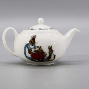 Può includere: Teiera in porcellana bianca con un design di Peter Rabbit. La teiera presenta una scena di Peter Rabbit e la sua famiglia. Il testo sulla teiera recita "The Very Busy Cottontail".
