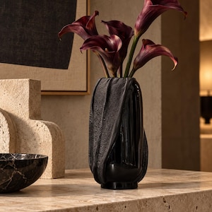 Op de afbeelding: Een hoge, glanzend zwarte vaas met een textuurrijke, stofachtige wikkel aan de buitenkant. De vaas bevat een boeket diepbordeauxrode calla-lelies met lange groene stelen. Het object heeft een gladde, reflecterende afwerking aan de onderkant.