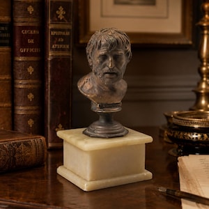 May include: A bronze-toned bust of a bearded man with short hair, mounted on a dark circular pedestal and a rectangular, off-white stone base. The sculpture features detailed facial features, including a furrowed brow and a slightly open mouth.