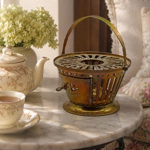 Puede incluir: Un calentador de té de latón con base circular, cuerpo cilíndrico de listones, puerta lateral abatible con un pequeño pestillo y una asa alta arqueada. La superficie superior presenta un diseño de recortes radiales.