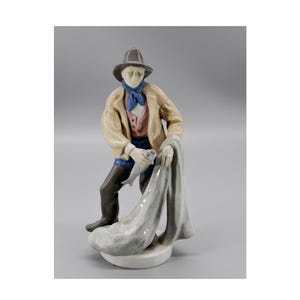 May include: A porcelain figurine of a fisherman holding a fish and a net. He is wearing a brown hat, a beige jacket, a blue shirt, and blue pants. The figurine is painted in realistic colors.