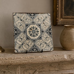 May include: A square ceramic tile featuring a dark blue and cream-colored intricate floral and geometric pattern. The design includes a central star-like flower motif surrounded by scrolling acanthus leaves and linear borders on a light background.