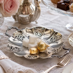 Può includere: Un piatto da portata in metallo argentato con due manici decorati e un bordo smerlato con intricati motivi traforati. Il piatto contiene tre zollette di zucchero bianche e due cioccolatini rotondi avvolti in carta stagnola dorata.