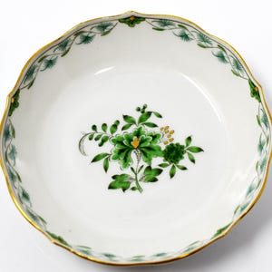 Könnte beinhalten: Eine weiße Porzellanschale mit gewelltem Rand und Goldrand. Die Schale hat ein zentrales Blumendesign in Grün- und Gelbtönen. Der Rand ist mit einem grünen Blättermuster verziert.