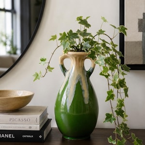 Könnte beinhalten: Eine Keramikvase mit zwei Henkeln, einer glänzend grünen Basis und einer cremefarbenen Tropfglasur am Hals. Die Vase enthält eine hängende Efeupflanze mit panaschierten grün-weißen Blättern, die einen kaskadierenden Effekt über die Seiten erzeugt.