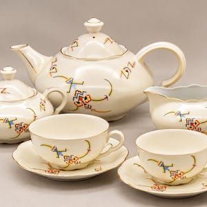 May include: A cream-colored porcelain tea set featuring a teapot with a lid, a lidded sugar bowl, a creamer, and two teacups with matching saucers. Each piece is decorated with a delicate, colorful geometric pattern and gold trim.