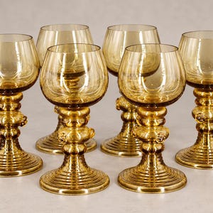 Può includere: Un set di cinque calici in vetro color ambra. Ogni calice presenta una coppa arrotondata, uno stelo con nodi di vetro decorativi in rilievo e un'ampia base circolare con una serie di anelli concentrici incisi sulla superficie del vetro.