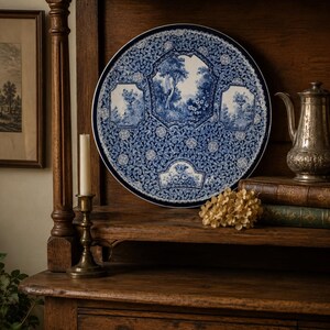 May include: A round ceramic plate featuring a dark blue and white transferware pattern. The design includes a central landscape scene with trees and flowers, surrounded by intricate floral motifs and smaller framed vignettes of nature scenes.