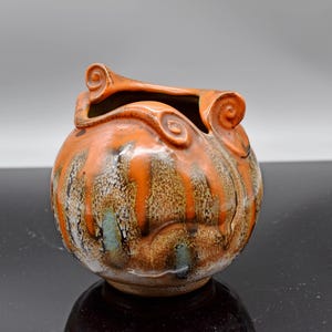 Może przedstawiać: Ręcznie wykonany ceramiczny wazon o okrągłym korpusie i otwartym wierzchu. Wazon ma żywy pomarańczowy szkliwo z pionowymi smugami brązu i bieli oraz dekoracyjnymi, zwiniętymi akcentami. Podstawa jest w kolorze ciemnobrązowym.
