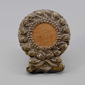 Puede incluir: Un marco de fotos plateado con un diseño de corona y un respaldo de madera. La corona está hecha de hojas y flores estilizadas.