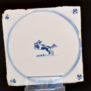 Peut inclure: Un carreau de céramique blanc avec un motif bleu et blanc d'un chien courant dans un cercle. Le carreau a une bordure bleue et quatre motifs de fleurs bleues dans les coins.