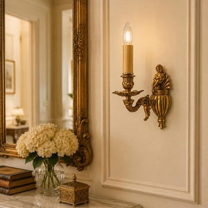 Può includere: Un'applique da parete color oro con un punto luce centrale a forma di candela e una lampadina a fiamma. La base in metallo è decorata con complessi rilievi, tra cui una piccola figura sulla sommità e una sezione inferiore scanalata a urna.