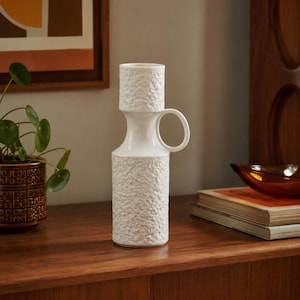 May include: A white ceramic vase with a cylindrical body and a smaller cylindrical top section, both featuring a textured, rough surface. The vase has a smooth, narrow neck and a single circular handle attached to the side of the upper section.
