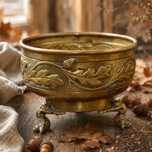 May include: A round brass bowl featuring an embossed design of oak leaves and acorns around the exterior. The bowl is supported by three ornate, claw-shaped feet and has a polished metallic finish with visible signs of age and patina.