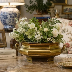 Peut inclure: Une jardinière en laiton poli de forme octogonale avec deux poignées latérales, remplie d'un arrangement luxuriant de fleurs blanches et de feuillage vert. Le récipient métallique présente un large bord et une base effilée.