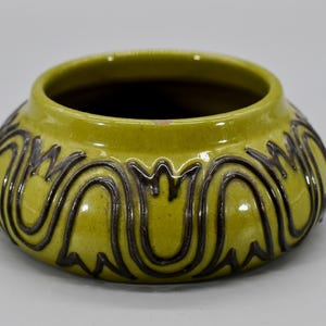 May include: A round, ceramic bowl in a vibrant chartreuse color. The bowl features a dark brown, stylized floral pattern around the exterior. The interior of the bowl is dark, creating a contrast with the bright exterior.