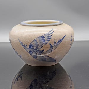Puede incluir: Un jarrón de cerámica color crema con un borde azul y un diseño de pájaro azul. El jarrón tiene forma redonda y una superficie craquelada. El pájaro está representado en vuelo con las alas extendidas.