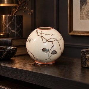 May include: A spherical ceramic vase with a cream-colored glaze, featuring hand-painted dark brown branches and small blue-grey leaves. The vase has a thin orange rim at the top opening and a matching orange base, creating a distinct contrast.