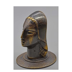May include: A bronze sculpture of a woman's head in profile. The head is stylized with a smooth surface and a detailed face. The sculpture has a dark bronze color with gold accents on the head covering and base.