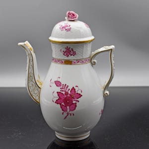 May include: White porcelain coffee pot with a pink floral design. The pot features a rose-shaped finial on the lid, gold trim, and a decorative handle. The floral pattern includes large, stylized flowers and smaller floral accents.