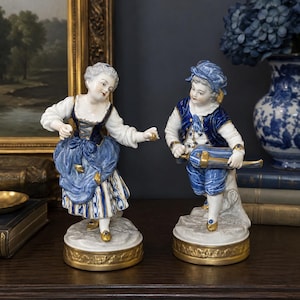 Może przedstawiać: Dwie porcelanowe figurki przedstawiające osobę w niebiesko-białej sukni oraz osobę w niebiesko-białym stroju grającą na instrumencie muzycznym. Obie postacie stoją na okrągłych podstawach ze złotym wykończeniem i mają szczegółowo malowane ubrania.