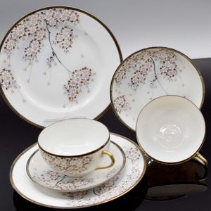 Könnte beinhalten: Weißes Porzellan-Teeservice mit floralem Kirschblüten-Design. Das Set beinhaltet einen großen Teller, einen kleineren Teller, eine Untertasse und eine Teetasse. Die Ränder und Griffe sind goldfarben.