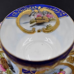 Könnte beinhalten: Ein Vintage-Teetassen- und Untertassen-Set mit blauem und goldenem Design. Tasse und Untertasse zeigen eine japanisch inspirierte Szene mit einem Haus, Bäumen und Blumen.