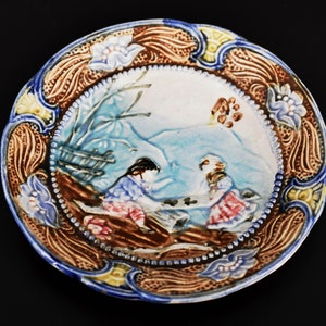 May include: A vintage ceramic plate with a raised design of two figures in a winter scene. The plate has a blue, brown, and white color scheme with floral accents.