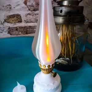 May include: A white ceramic oil lamp with a gold-colored base and a white frosted glass shade. The lamp is lit and has a warm orange glow. The lamp is sitting on a blue surface.