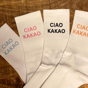 Könnte beinhalten: Vier weiße Socken mit dem Schriftzug "Ciao Kakao" in verschiedenen Farben: hellblau, rosa, schwarz und orange.