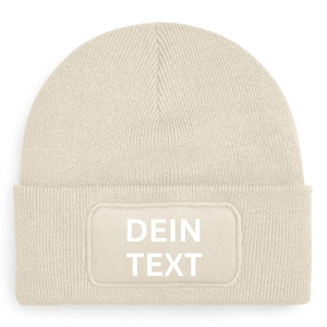Könnte beinhalten: Eine beige Strickmütze mit einem weißen Patch, auf dem "DEIN TEXT" steht.