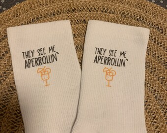 May include: A pair of white socks with the text "THEY SEE ME APERROLLIN'" printed in black. Each sock features a small orange cocktail graphic. The socks are displayed on a woven, tan surface.