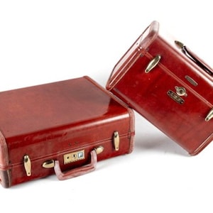 May include: Two rectangular, deep red suitcases with a glossy finish. Each case features gold-toned metal latches, a sturdy handle, and a central locking mechanism. The left suitcase displays a small square plate with the letter F on the lock.