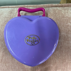 Op de afbeelding: Een paars hartvormig Polly Pocket speelgoed met een roze handvat. Het speelgoed heeft een gouden logo met de tekst "Polly Pocket" en een hart.