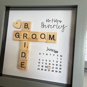 Peut inclure: Une impression encadrée avec des carreaux en bois qui épellent "Bride & Groom" avec un cœur sur le carreau "B". L'impression comprend également un calendrier pour juin 2024 avec un cœur sur le 21. Le texte "Mr & Mrs Beverley" est écrit en cursive en haut de l'impression.