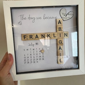 Könnte beinhalten: Ein gerahmtes personalisiertes Kunstwerk mit den Worten "The day we became" in eleganter Schrift. Der Name "Franklin" ist mit Holzbuchstaben geschrieben, gekreuzt mit "Abigail". Ein Kalender hebt den Juli 2025 hervor, mit einem Herz, das ein Datum markiert.
