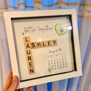 Puede incluir: Un marco cuadrado blanco tipo caja con una pantalla personalizada que incluye el texto Better Together, un recorte de mapa en forma de corazón, fichas de letras de madera que forman LAUREN y ASHLEY, y un calendario de agosto de 2025 con un corazón.