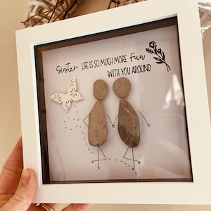 May include: A framed art piece with a white border. The artwork features two stone figures holding hands, a butterfly, and the text "Sister life is so much more fun with you around."