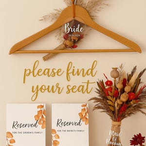 Può includere: Una gruccia di legno con un cartello Bride in acrilico trasparente e la data 24.06.2024. Le parole "please find your seat" sono in oro. Due cartelli "Reserved" e un bouquet di fiori secchi completano l'arredamento del matrimonio.