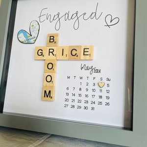 Puede incluir: Un anuncio de compromiso enmarcado con un calendario de madera que muestra la fecha del 11 de mayo de 2024. La palabra "Engaged" está escrita en cursiva con un símbolo de corazón. La palabra "GROOM" está deletreada con fichas de letras de madera, con la letra "O" apareciendo dos veces.