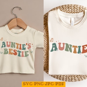 May include: Two cream-colored t-shirts with a retro-style font design. The smaller shirt says "Auntie's Bestie" with colorful letters and flowers. The larger shirt says "Auntie" with colorful letters and flowers.